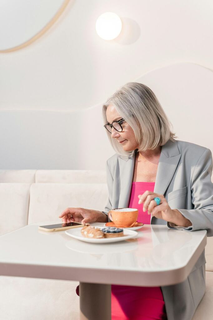 Senior woman in a gray blazer enjoying coffee and pastries at a table with a phone.